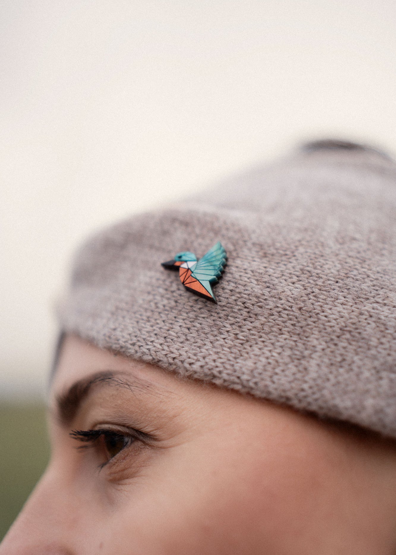 A close-up of a person wearing a knit beige hat decorated with a colorful kingfisher pin in shades of teal, blue, and orange. This eco-friendly accessory is perfect for bird lovers. The person's eyebrow and eye are partially visible.