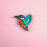 A colorful, geometric kingfisher pin brooch shaped like a bird with turquoise wings and head, and an orange body, handcrafted from recycled wood, set against a solid pink background.