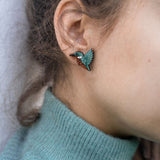 Close-up of a person wearing a turquoise sweater and eco-friendly Kingfisher Stud Earrings, shaped like a hummingbird, with the focus on their ear and earring. The background is softly blurred.