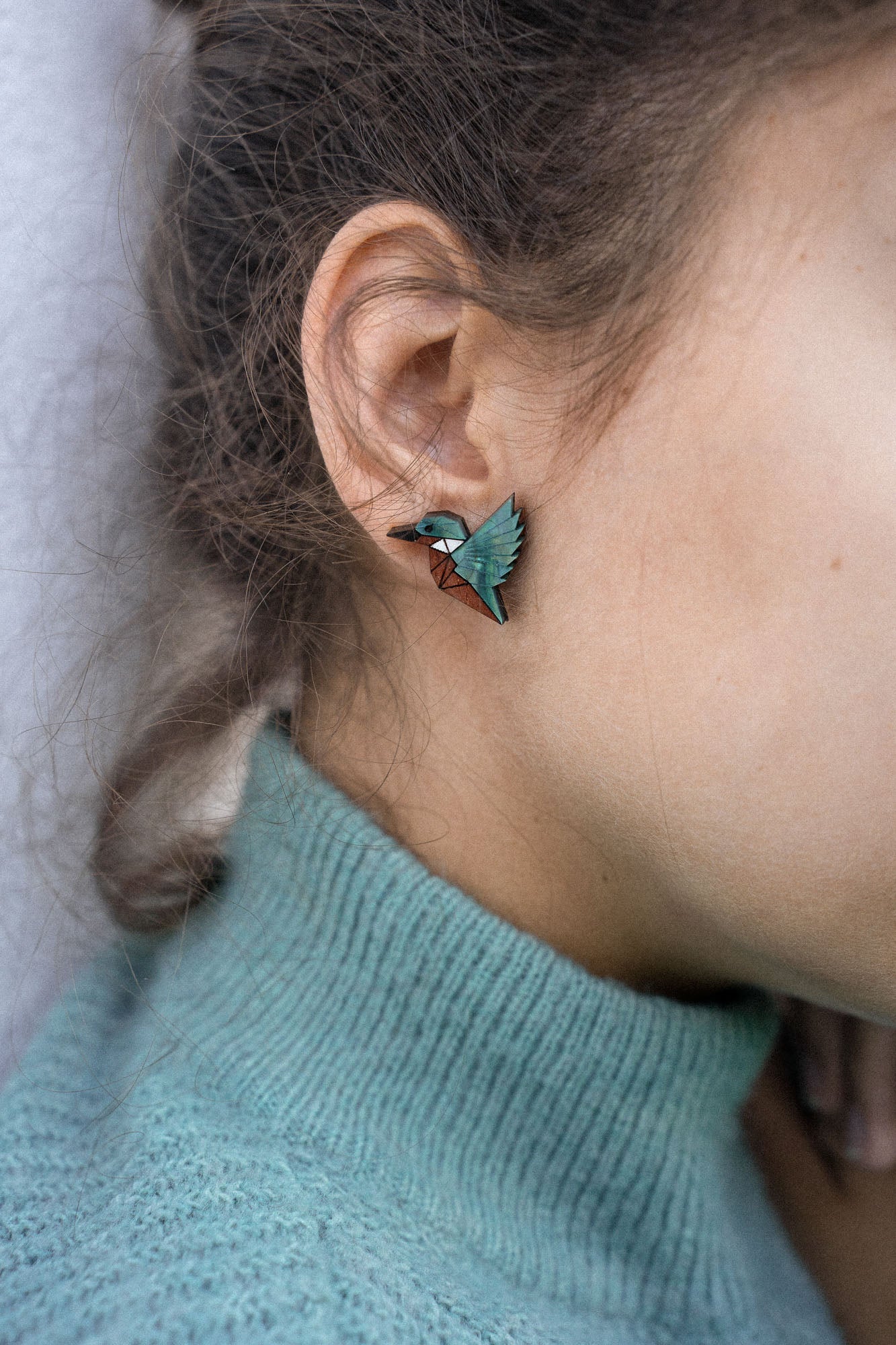 Close-up of a person wearing a turquoise sweater and eco-friendly Kingfisher Stud Earrings, shaped like a hummingbird, with the focus on their ear and earring. The background is softly blurred.