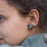 Close-up of a person wearing a turquoise sweater and colorful, sustainably made hummingbird-shaped earring, with only part of their face and ear visible.