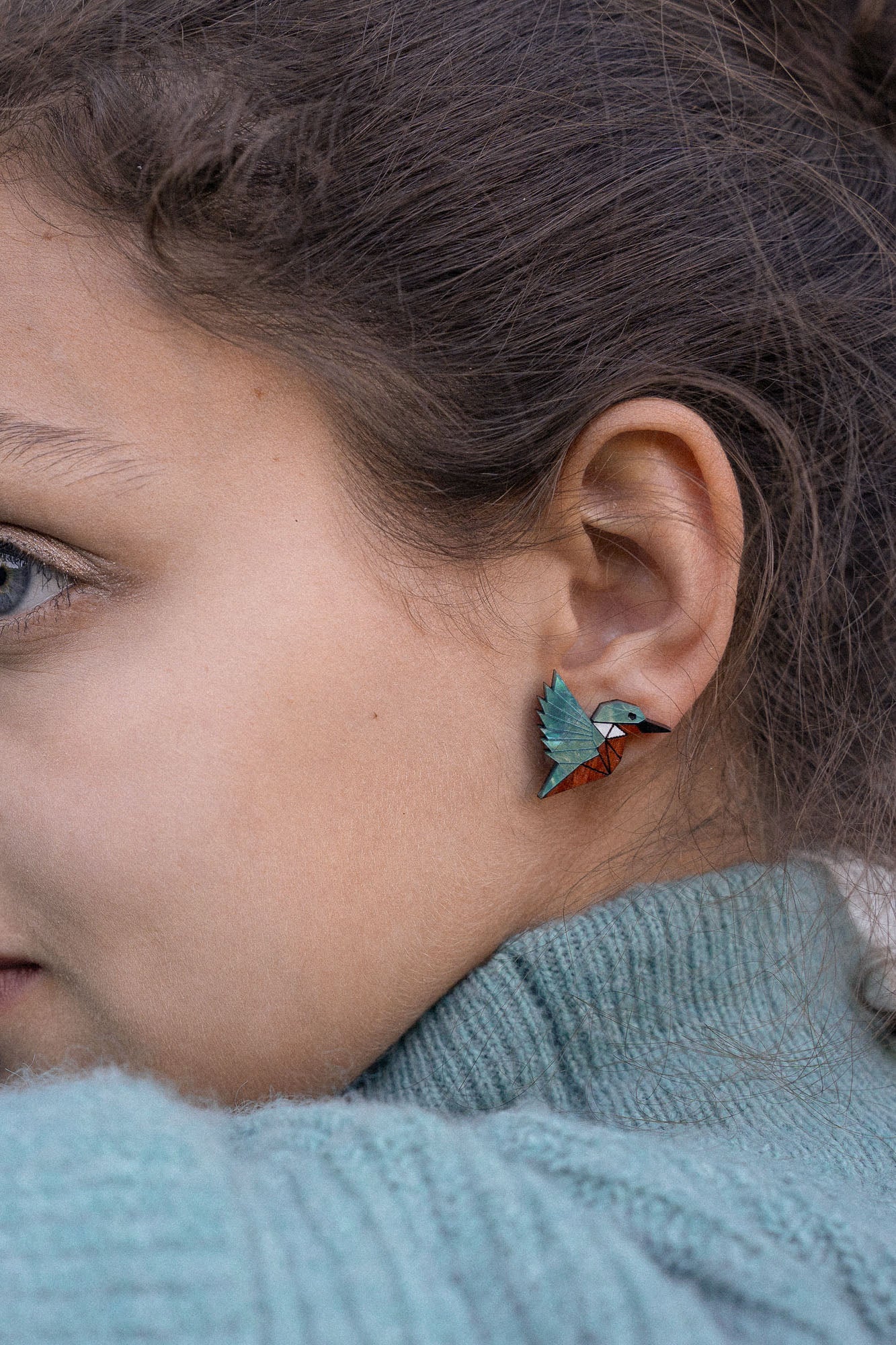 Close-up of a person wearing a turquoise sweater and colorful, sustainably made hummingbird-shaped earring, with only part of their face and ear visible.