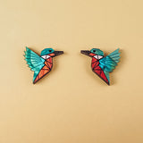 Two colorful, geometric hummingbird brooches with turquoise wings and red bodies face each other on a plain beige background, each piece expertly crafted to be eco-friendly and sustainably made.