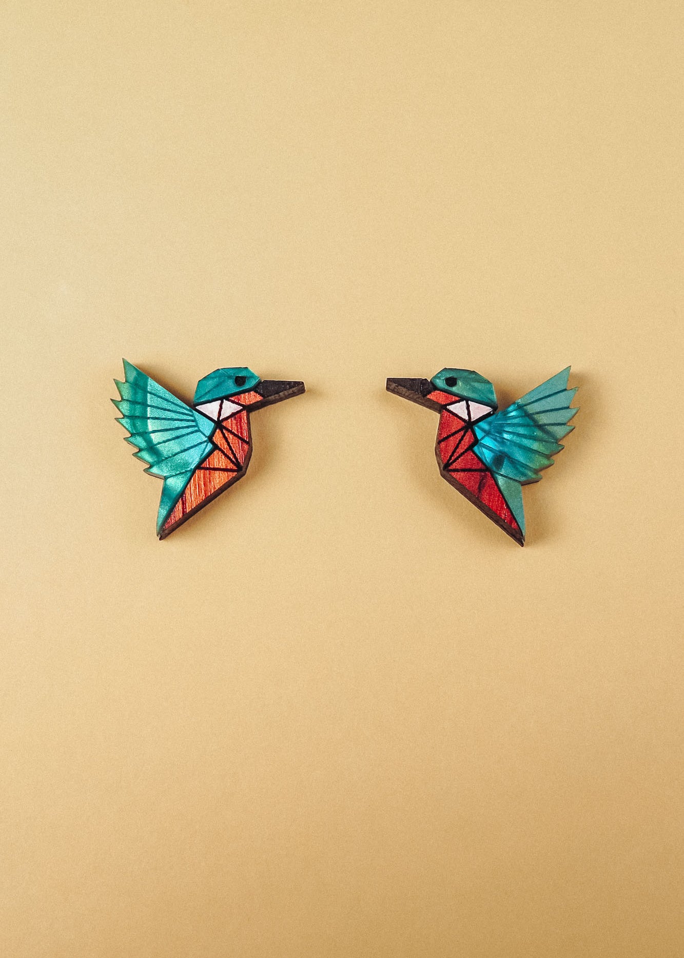 Two colorful, geometric hummingbird brooches with turquoise wings and red bodies face each other on a plain beige background, each piece expertly crafted to be eco-friendly and sustainably made.