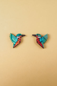 Two colorful, geometric hummingbird brooches with turquoise wings and red bodies face each other on a plain beige background, each piece expertly crafted to be eco-friendly and sustainably made.