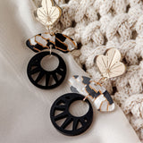 A pair of modern, handcrafted Kintsugi Earrings with gold floral tops, marbled oval shapes, and black circular cutouts, displayed on white fabric next to a textured, cream knitted cloth.