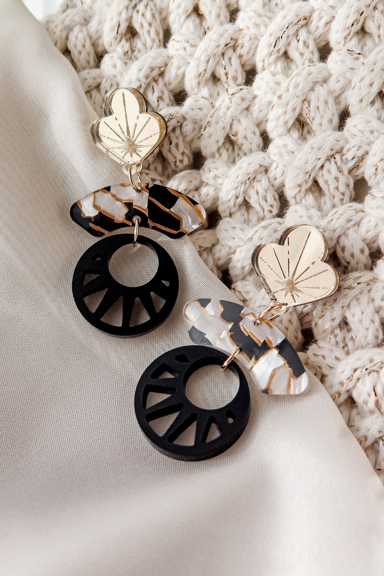 A pair of modern, handcrafted Kintsugi Earrings with gold floral tops, marbled oval shapes, and black circular cutouts, displayed on white fabric next to a textured, cream knitted cloth.