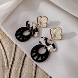 A pair of stylish Kintsugi Earrings featuring gold flower-shaped studs, marbled white and brown connectors, and large black circular pendants, all handcrafted and displayed on a textured beige fabric background.