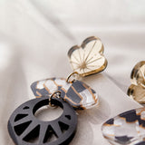 Close-up of handcrafted Kintsugi Earrings, featuring gold heart-shaped tops, marbled white and brown oval centers, and black circular pendants with geometric cutouts, displayed on a soft, light-colored fabric background.