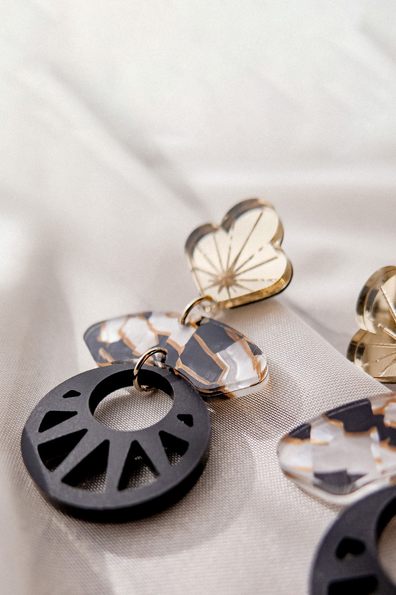 Close-up of handcrafted Kintsugi Earrings, featuring gold heart-shaped tops, marbled white and brown oval centers, and black circular pendants with geometric cutouts, displayed on a soft, light-colored fabric background.