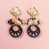 A pair of handcrafted statement earrings on a pink background, featuring gold flower-shaped tops, marbled tortoiseshell-patterned bars, and large black circular pendants with cutout details inspired by Japanese art.