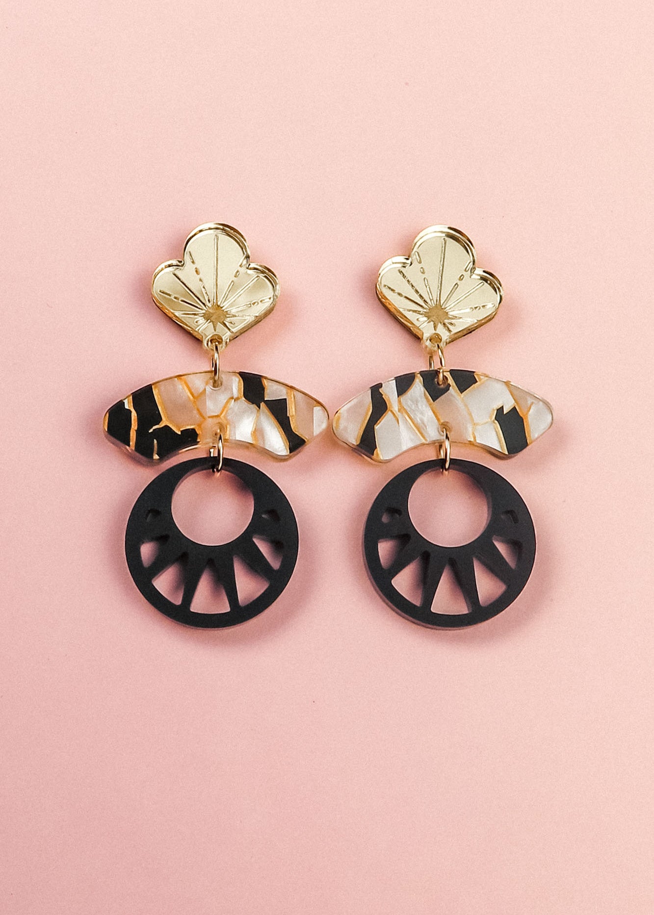 A pair of handcrafted statement earrings on a pink background, featuring gold flower-shaped tops, marbled tortoiseshell-patterned bars, and large black circular pendants with cutout details inspired by Japanese art.