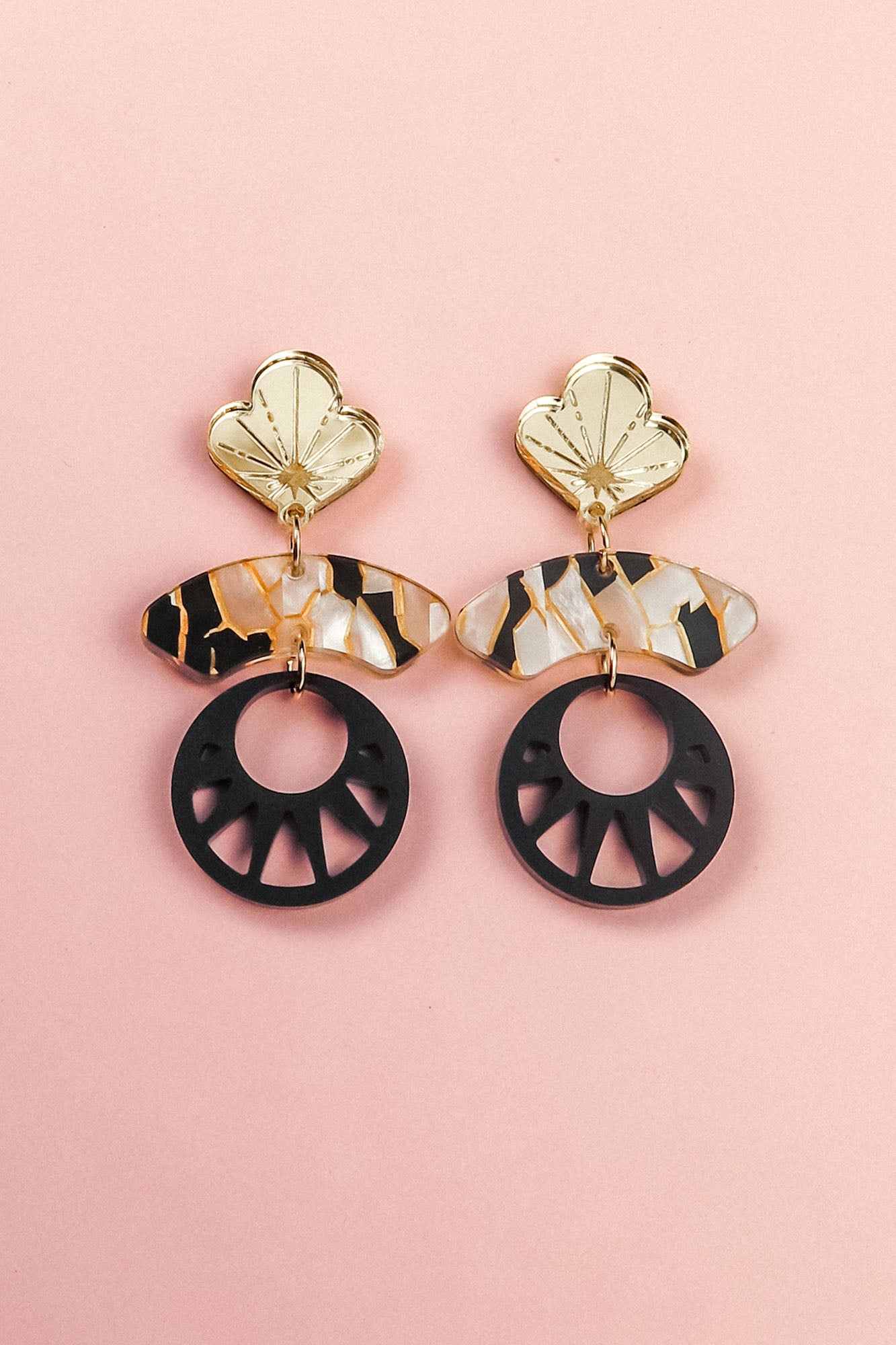 A pair of handcrafted statement earrings on a pink background, featuring gold flower-shaped tops, marbled tortoiseshell-patterned bars, and large black circular pendants with cutout details inspired by Japanese art.