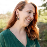 A woman with long auburn hair, wearing a green dress and handcrafted Kintsugi Earrings, smiles while looking down. She is outdoors with greenery and soft sunlight in the background.