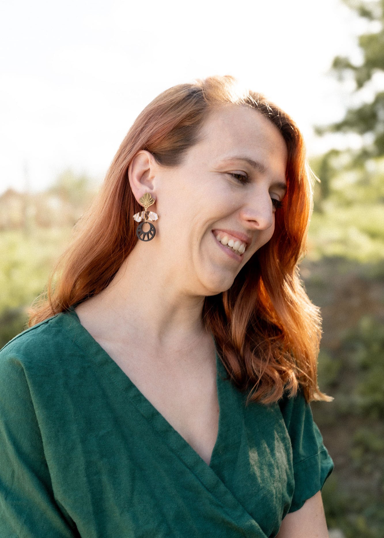 A woman with long auburn hair, wearing a green dress and handcrafted Kintsugi Earrings, smiles while looking down. She is outdoors with greenery and soft sunlight in the background.