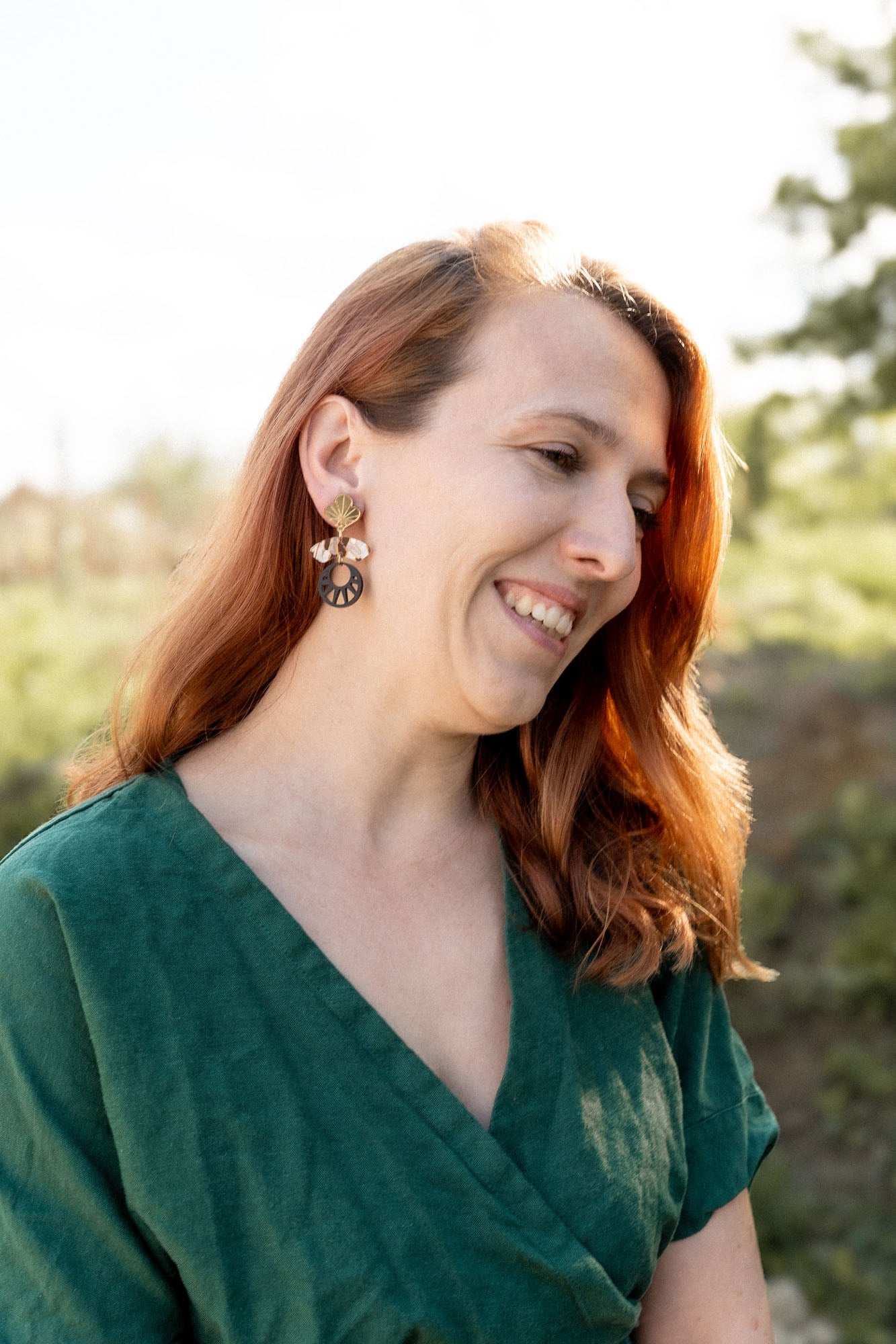 A woman with long auburn hair, wearing a green dress and handcrafted Kintsugi Earrings, smiles while looking down. She is outdoors with greenery and soft sunlight in the background.