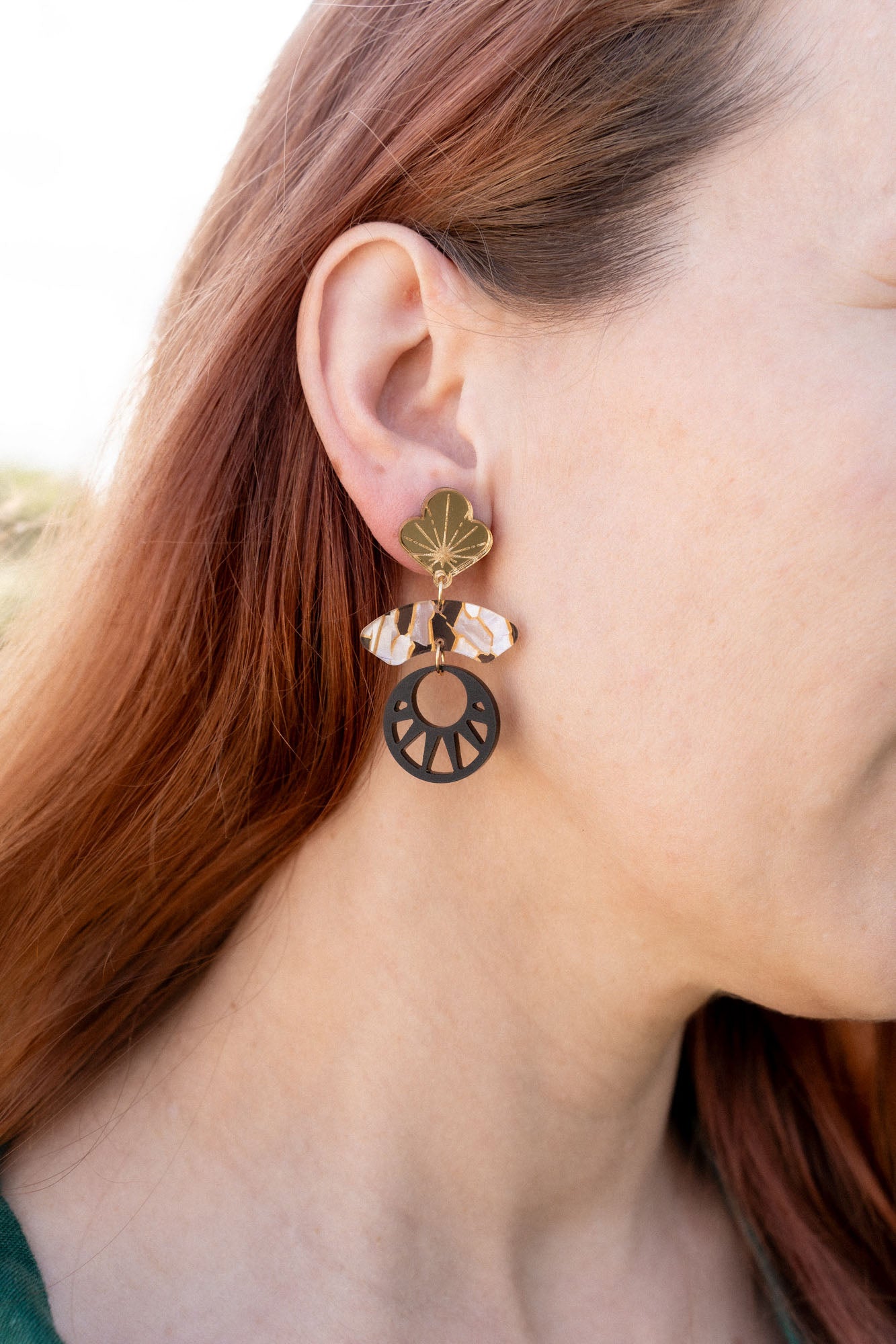 Close-up of a person with long auburn hair wearing handcrafted Kintsugi Earrings featuring a gold leaf, a clear crystal, and a black circular charm with a cut-out sunburst design inspired by Japanese art.