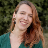 A woman with long, reddish-brown hair smiles warmly at the camera. She is wearing a green top and striking handcrafted Kintsugi Earrings. The blurred greenery in the background suggests an outdoor setting.