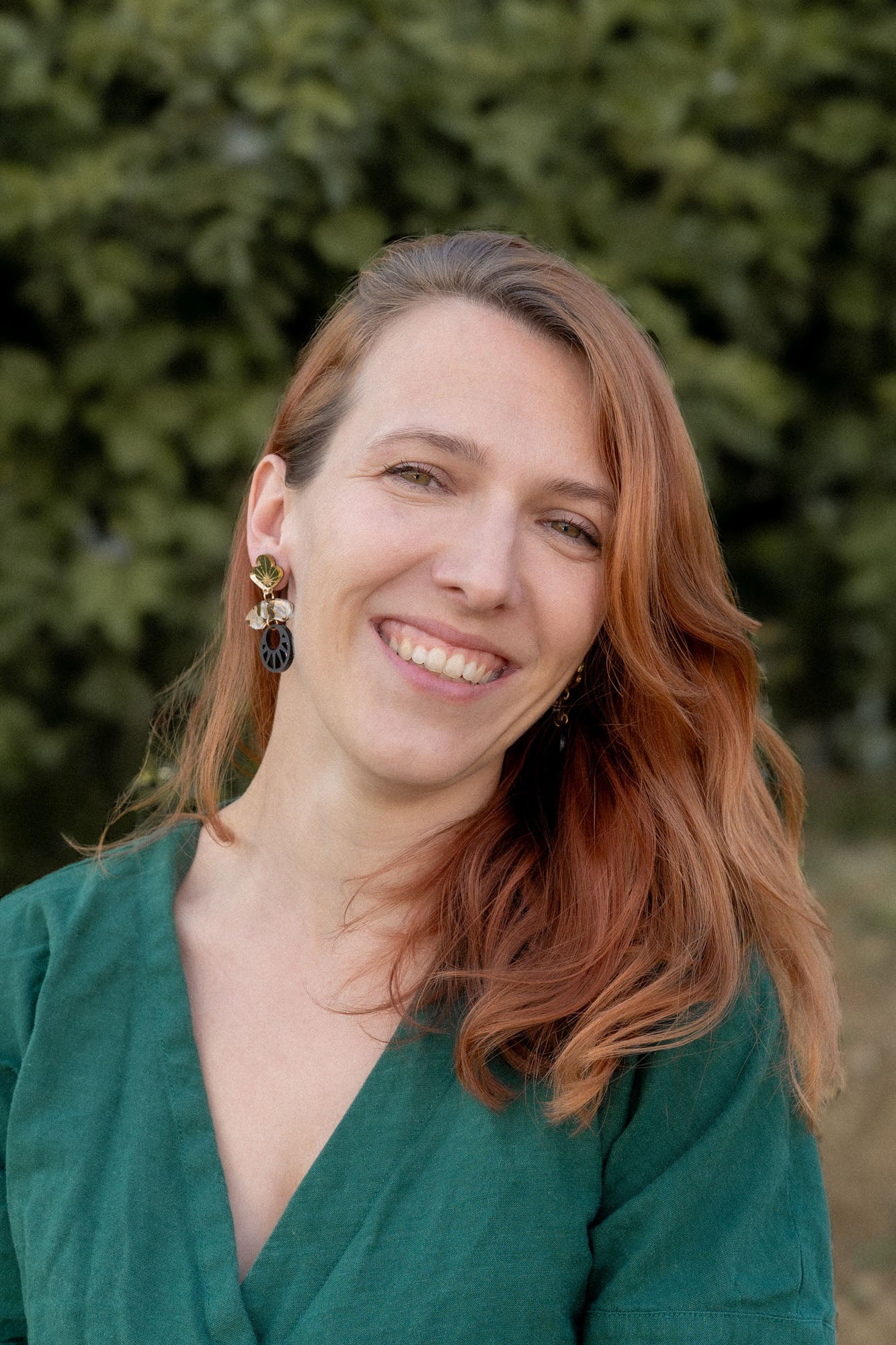 A woman with long, reddish-brown hair smiles warmly at the camera. She is wearing a green top and striking handcrafted Kintsugi Earrings. The blurred greenery in the background suggests an outdoor setting.