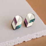 A pair of geometric stud earrings with silver outlines, featuring sections of teal, purple, and cream enamel, perfect for lovers of minimalist jewelry and displayed on a textured white and brown surface.