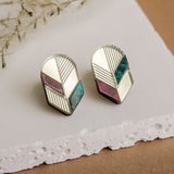 A pair of geometric stud earrings featuring silver edges and sections of white, mauve, and teal—beautifully displayed on a white textured slab with a brown background and dried plant decor—perfect for lovers of minimalist jewelry.