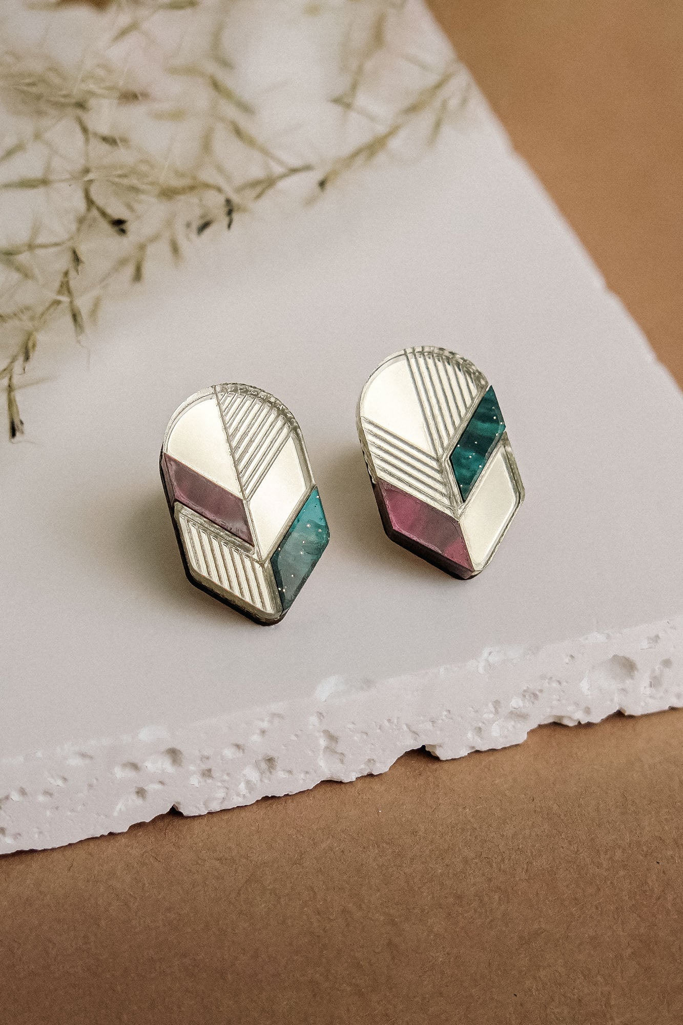 A pair of geometric stud earrings featuring silver edges and sections of white, mauve, and teal—beautifully displayed on a white textured slab with a brown background and dried plant decor—perfect for lovers of minimalist jewelry.