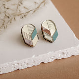 A pair of handmade geometric stud earrings with gold, pink, white, and teal sections, displayed on a white textured slab against a beige background with dried botanical accents—perfect for fans of minimalist jewelry.