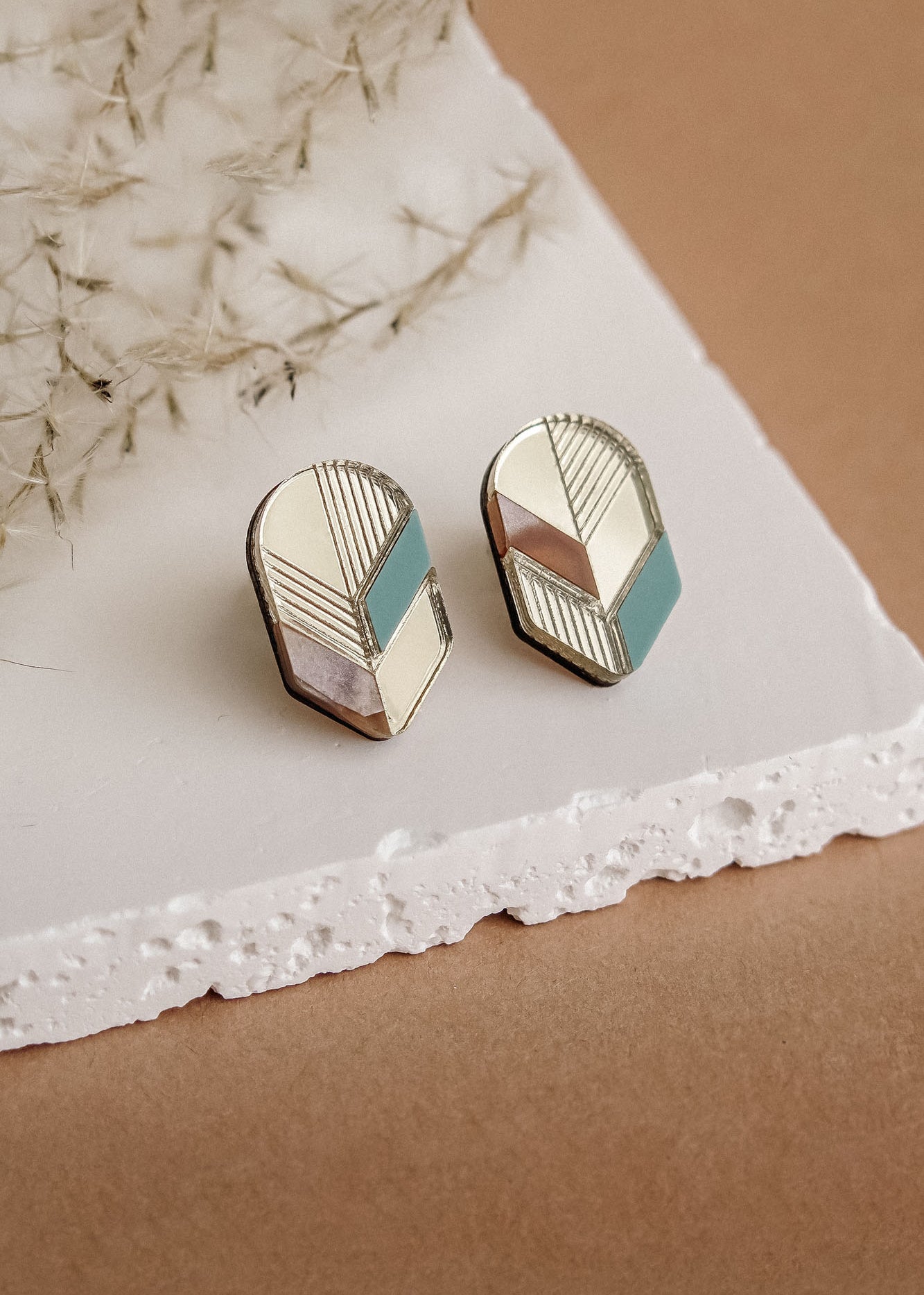 A pair of handmade geometric stud earrings with gold, pink, white, and teal sections, displayed on a white textured slab against a beige background with dried botanical accents—perfect for fans of minimalist jewelry.