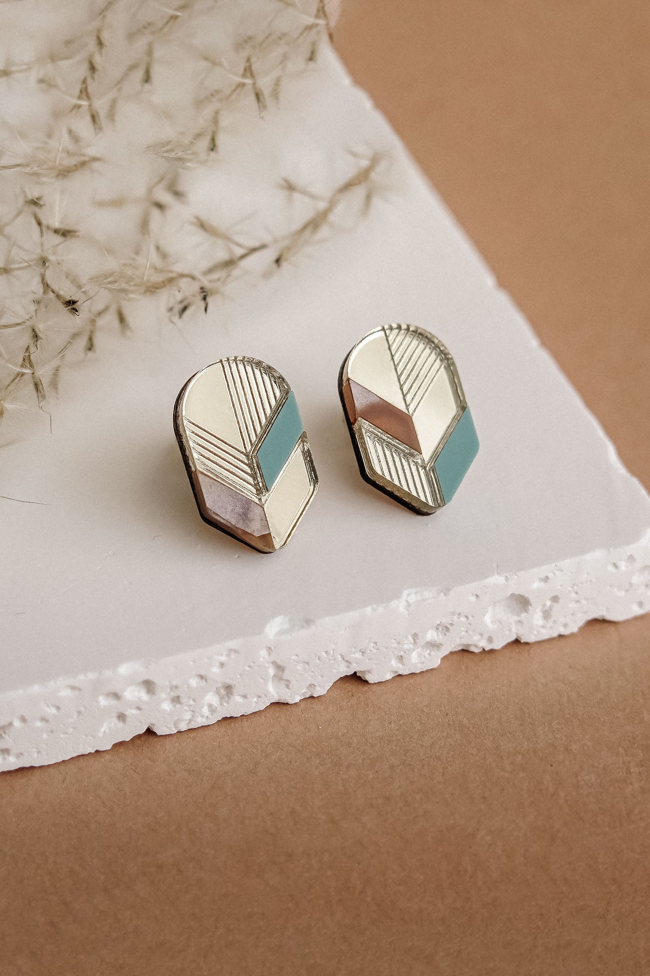 A pair of handmade geometric stud earrings with gold, pink, white, and teal sections, displayed on a white textured slab against a beige background with dried botanical accents—perfect for fans of minimalist jewelry.