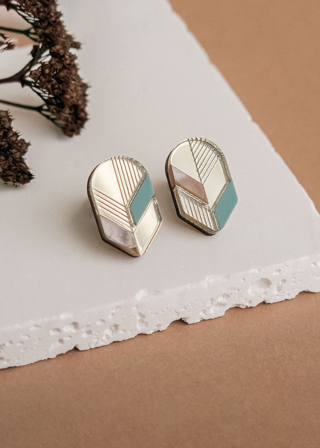 Handmade geometric earrings with gold, white, beige, and teal sections displayed on a white textured surface, dried flowers in the background—perfect for lovers of minimalist jewelry.