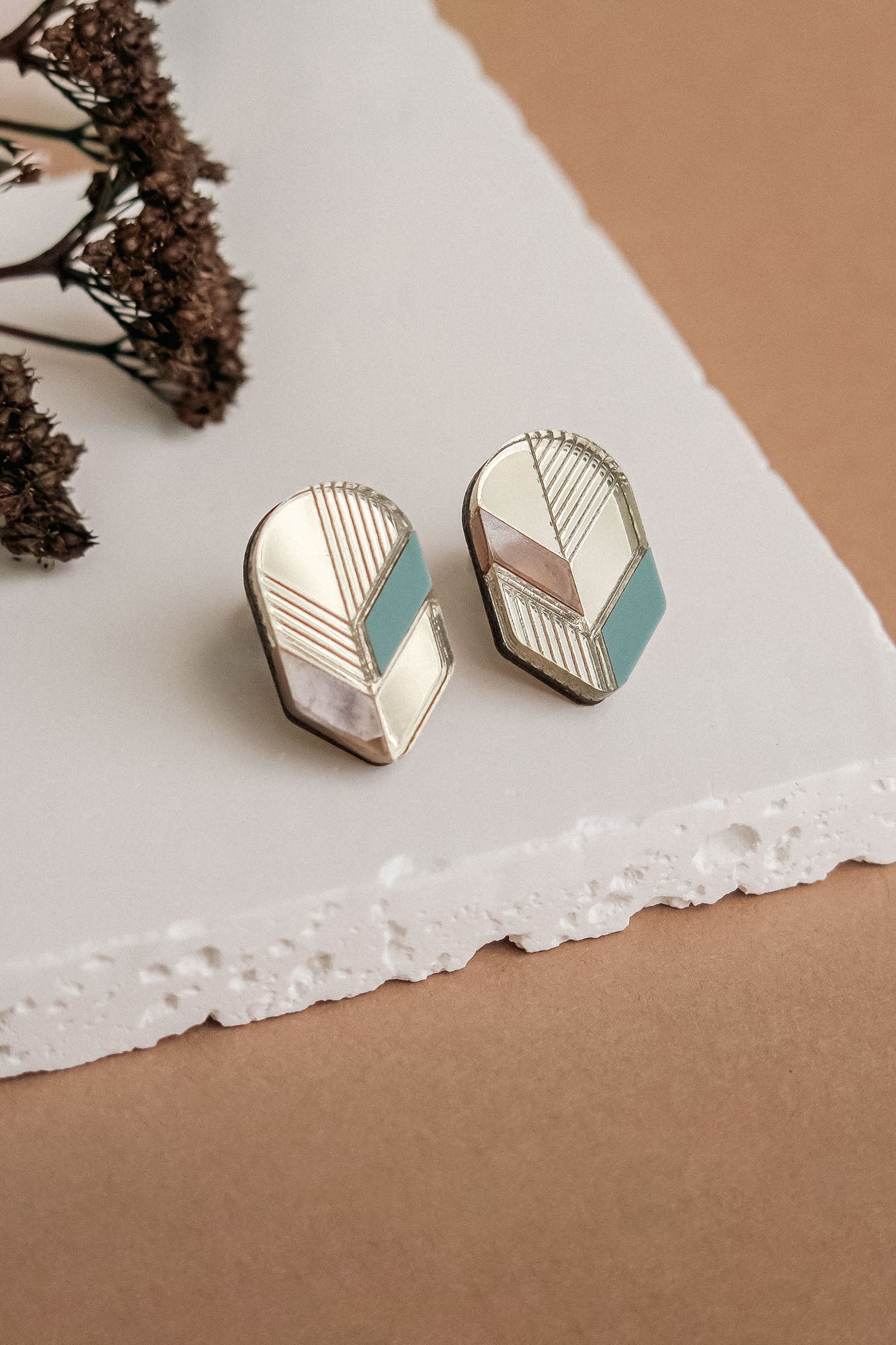 Handmade geometric earrings with gold, white, beige, and teal sections displayed on a white textured surface, dried flowers in the background—perfect for lovers of minimalist jewelry.