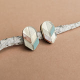 A pair of geometric Leaf Studs in gold, silver, and teal rest on a textured white branch against a light brown background—perfect for lovers of minimalist jewelry.