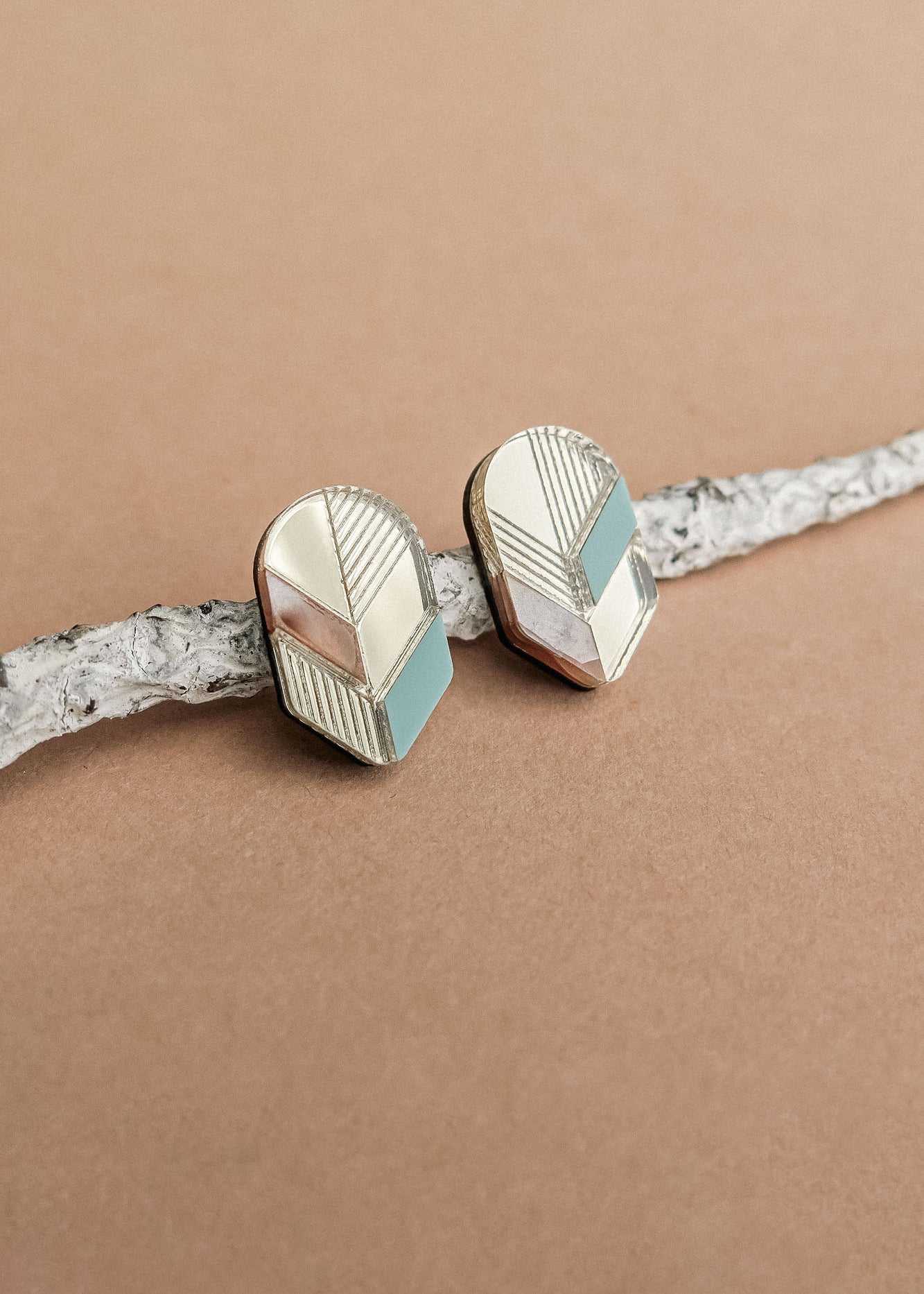 A pair of geometric Leaf Studs in gold, silver, and teal rest on a textured white branch against a light brown background—perfect for lovers of minimalist jewelry.