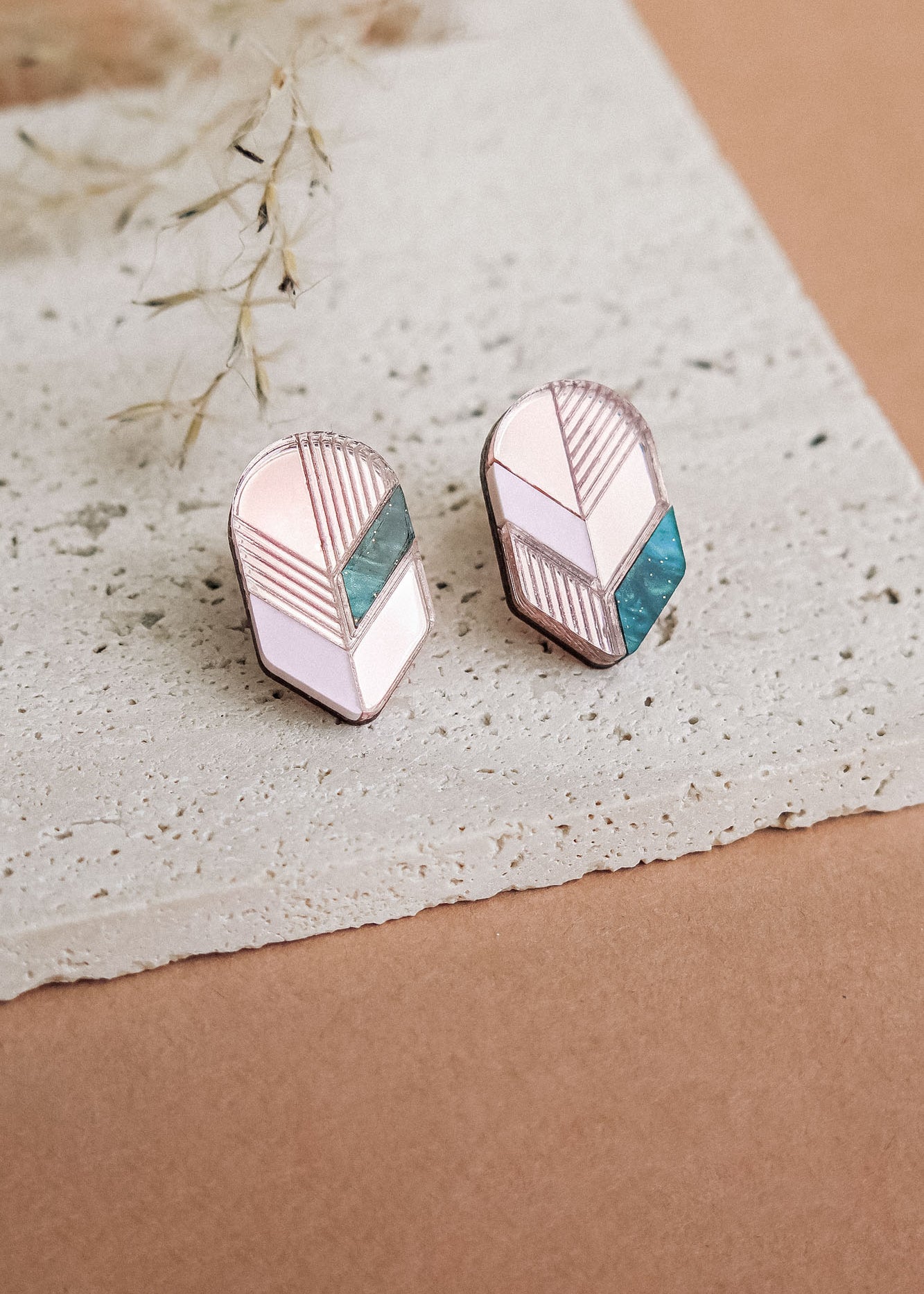 A pair of geometric stud earrings with pink, white, and teal segments set in silver, displayed on a textured white stone surface with dried grass in the background—perfect for lovers of minimalist jewelry.