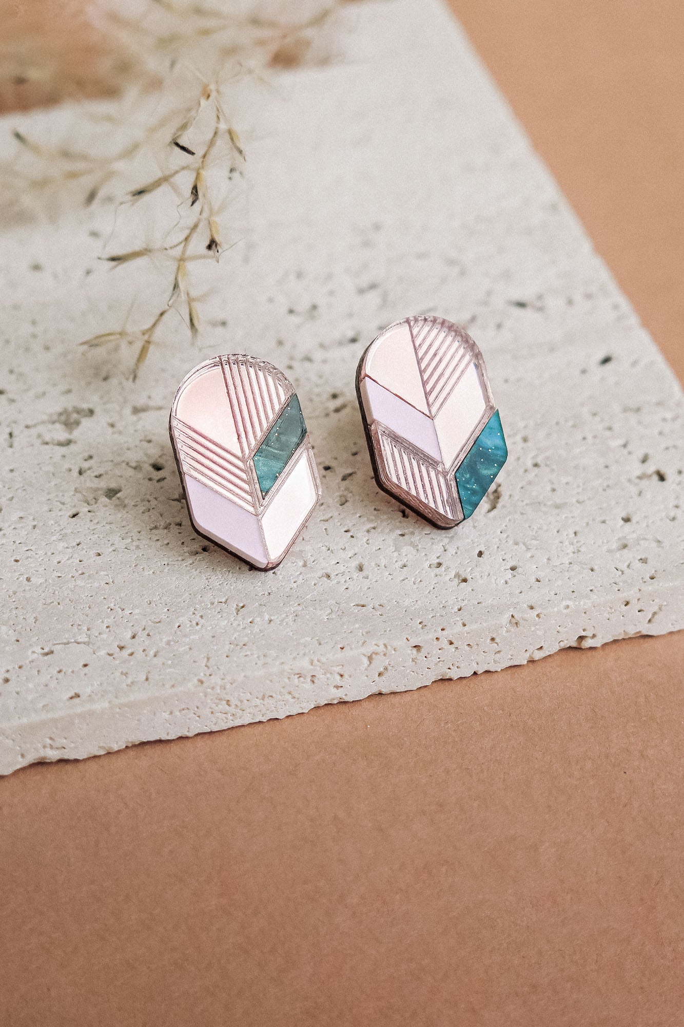 A pair of geometric stud earrings with pink, white, and teal segments set in silver, displayed on a textured white stone surface with dried grass in the background—perfect for lovers of minimalist jewelry.