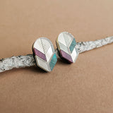 Handmade geometric stud earrings with silver, lavender, and teal sections rest on a textured, light gray branch against a beige background—a perfect touch of minimalist jewelry.