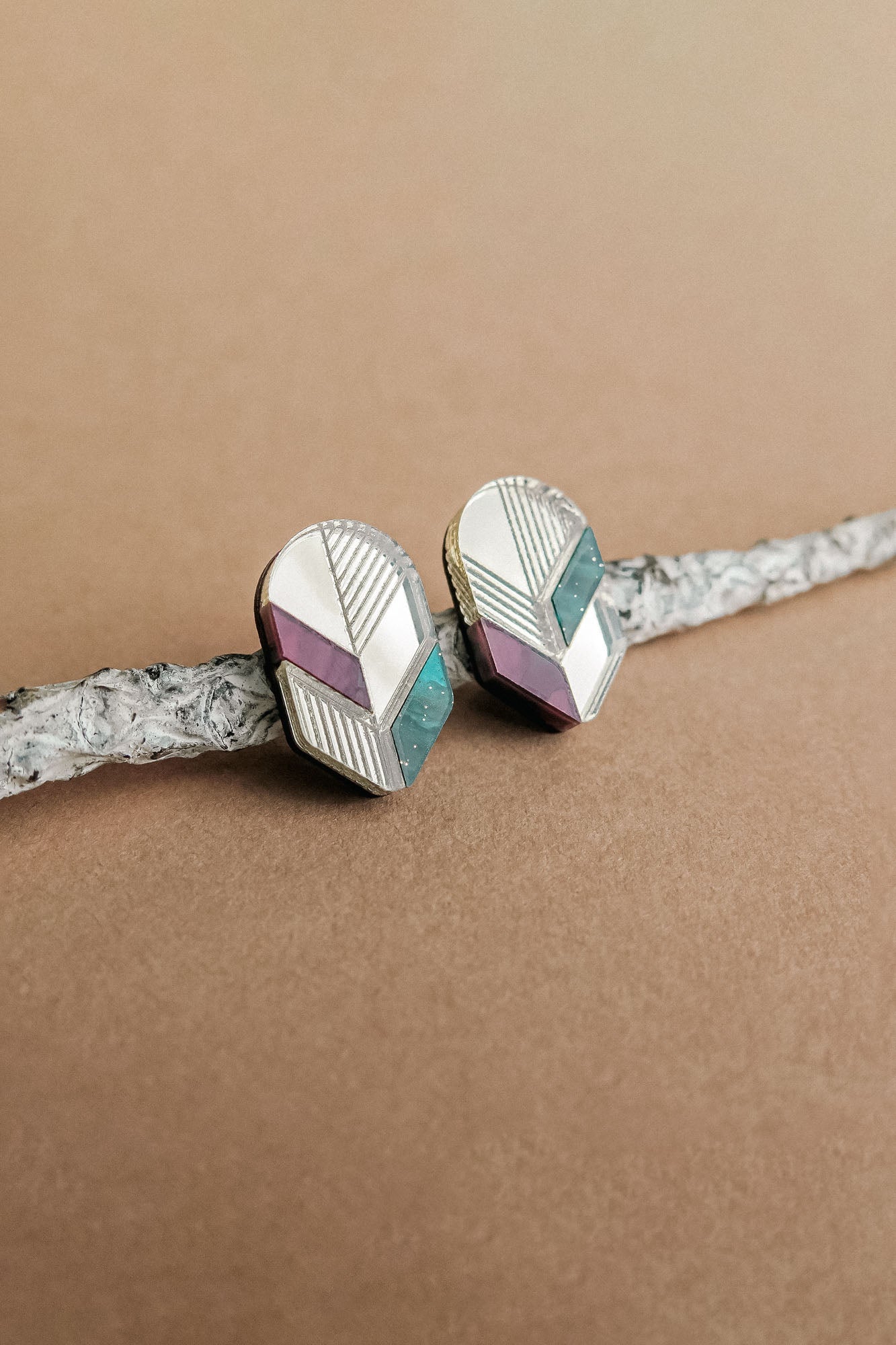 Handmade geometric stud earrings with silver, lavender, and teal sections rest on a textured, light gray branch against a beige background—a perfect touch of minimalist jewelry.