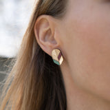Close-up of a womans ear wearing a geometric, gold-toned stud earring with pastel accents. This piece of minimalist jewelry stands out against the softly blurred background, highlighting its unique handmade design and part of her hair and face.