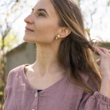 A woman with long brown hair wearing a mauve blouse and minimalist jewelry—heart-shaped earrings—looks upward while gently touching her hair; soft natural light and a blurred outdoor background.