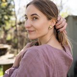 A woman with light brown hair in a low ponytail stands outdoors, wearing a mauve blouse and minimalist jewelry—gold leaf studs that catch the light. She looks over her shoulder at the camera, smiling softly, with one hand touching her hair.