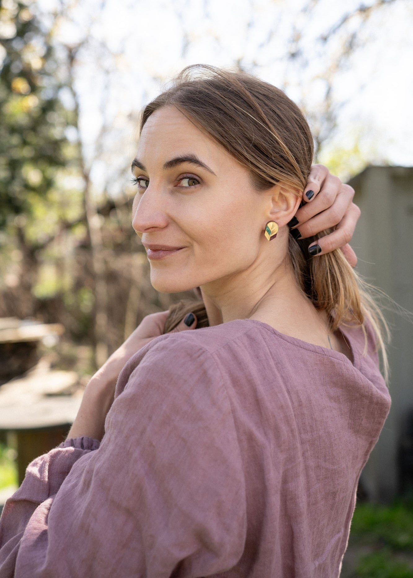 A woman with light brown hair in a low ponytail stands outdoors, wearing a mauve blouse and minimalist jewelry—gold leaf studs that catch the light. She looks over her shoulder at the camera, smiling softly, with one hand touching her hair.