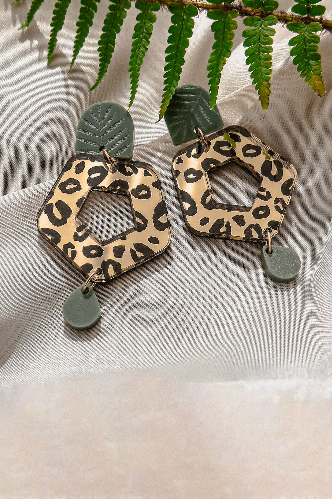 A pair of Leopard Earrings featuring timeless patterns—leopard print hexagons with green leaf-like and circular accents—crafted from premium materials, displayed on light fabric with a fern leaf above.