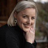 A woman with short blonde hair wearing a black sweater and striking Leopard Earrings smiles warmly while resting her chin on her hand. The background is softly blurred with natural, outdoor elements.