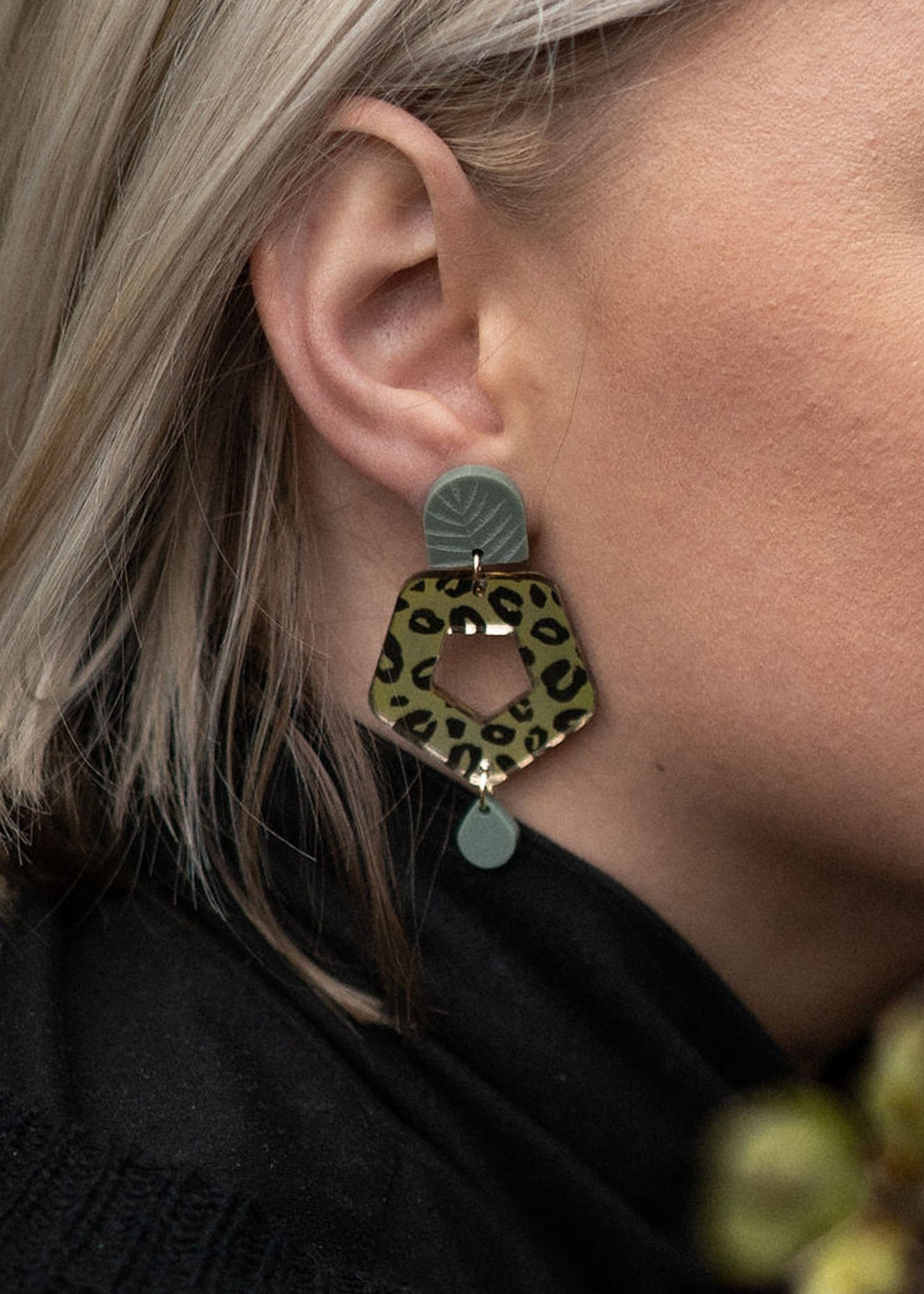 Close-up of a person with light blond hair wearing Leopard Earrings crafted from premium materials, featuring a leopard print hexagonal shape with green and gold accents for an eye-catching look.
