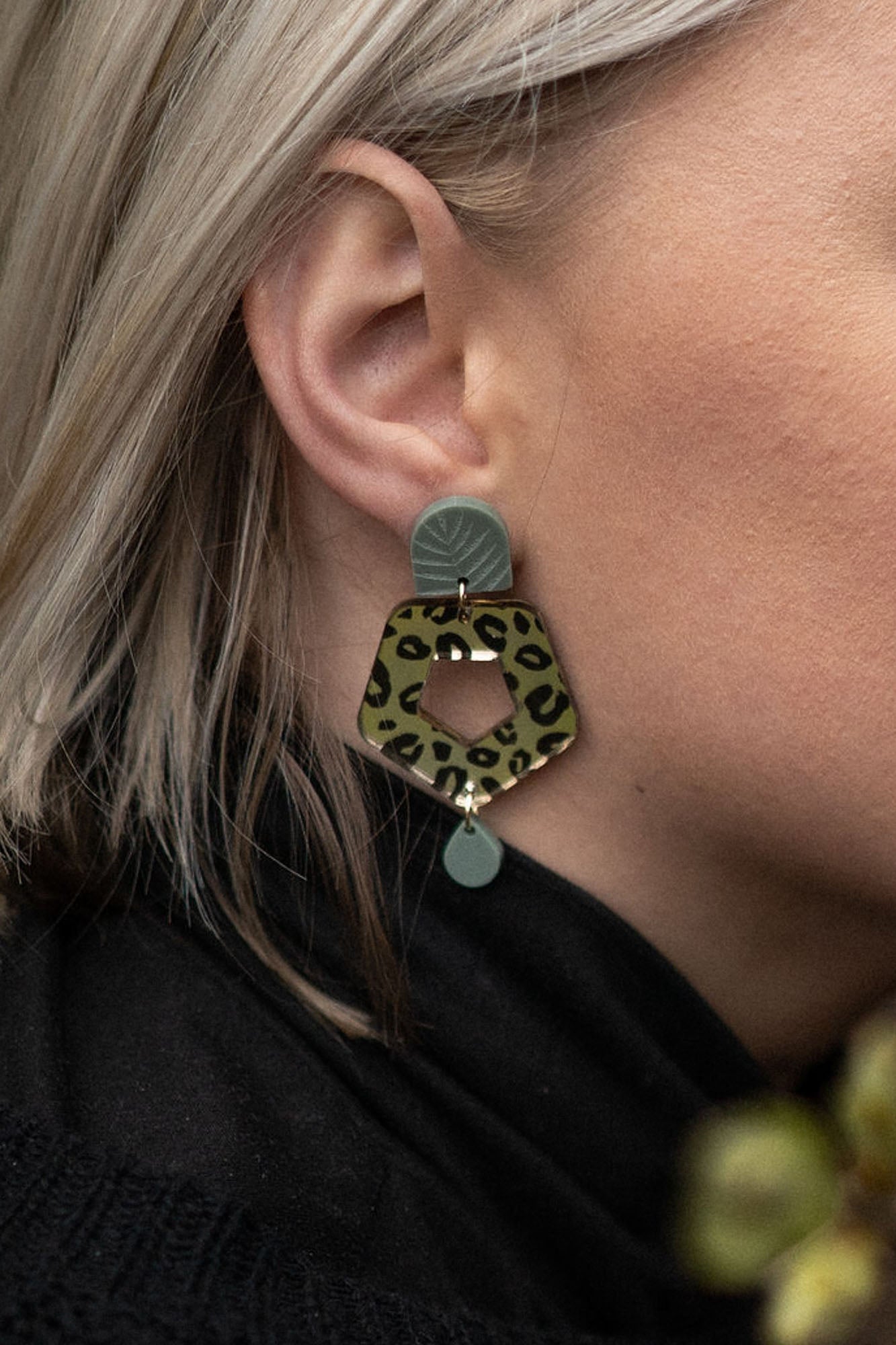 Close-up of a person with light blond hair wearing Leopard Earrings crafted from premium materials, featuring a leopard print hexagonal shape with green and gold accents for an eye-catching look.