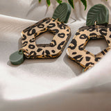 A pair of Leopard Earrings with timeless patterns—featuring leopard print hexagons and green leaf-shaped accents—rests on white satin fabric, with a fern leaf partially visible above, showcasing premium materials and unique style.