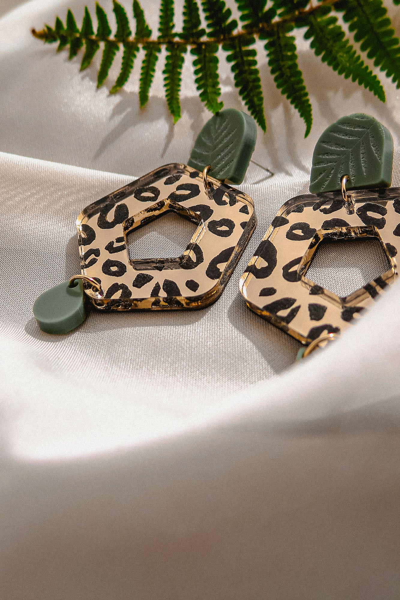 A pair of Leopard Earrings with timeless patterns—featuring leopard print hexagons and green leaf-shaped accents—rests on white satin fabric, with a fern leaf partially visible above, showcasing premium materials and unique style.
