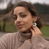 A woman with brown hair in a loose bun wears a beige turtleneck sweater and holds a large Leopard Earring to her ear. She is outdoors, with a blurred background of trees and grass, highlighting the allure of timeless patterns.