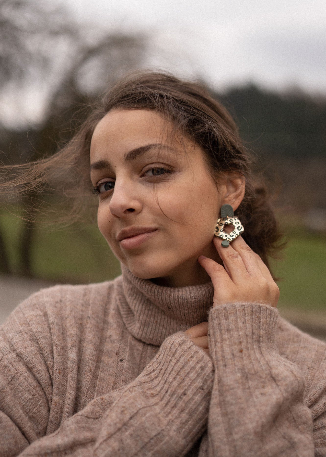 A woman with brown hair in a loose bun wears a beige turtleneck sweater and holds a large Leopard Earring to her ear. She is outdoors, with a blurred background of trees and grass, highlighting the allure of timeless patterns.