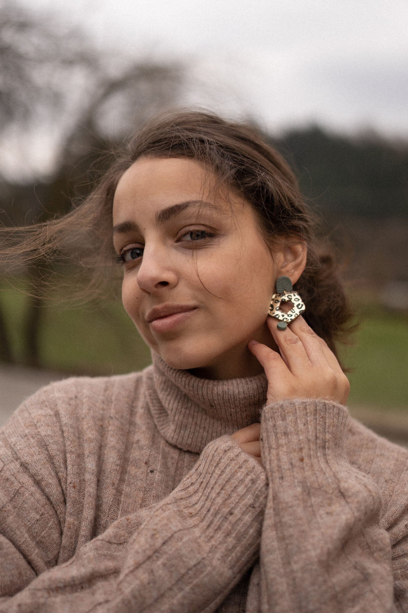 A woman with brown hair in a loose bun wears a beige turtleneck sweater and holds a large Leopard Earring to her ear. She is outdoors, with a blurred background of trees and grass, highlighting the allure of timeless patterns.
